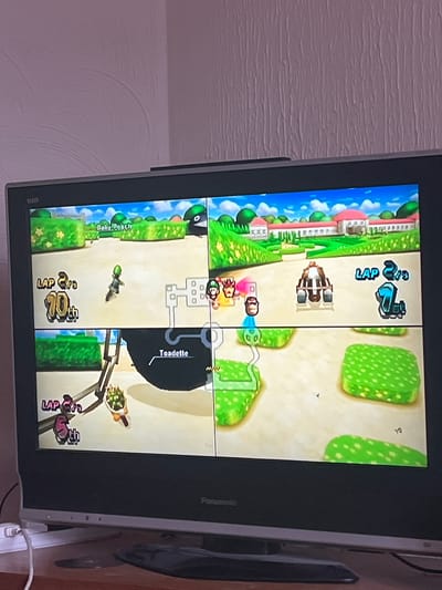 The image shows a television screen displaying the gameplay of Mario Kart Wii, likely recorded from a home in Haddenham, United Kingdom. The screen is divided into four sections, each showing a different player's perspective of the same race. The top left displays a racer on a course with blue skies, trees, and a white fence. The top right shows the view of a racer on a sandy track with green patches. The bottom left is a view of "Baby Beach" and a racer. The bottom right displays a racer near a wooden structure. The name "Toadette" is visible on the screen. The race is on its second lap. The television is a Panasonic brand. A wall is visible on the left side of the television. A table is seen on the right. A white cable is plugged into the TV.