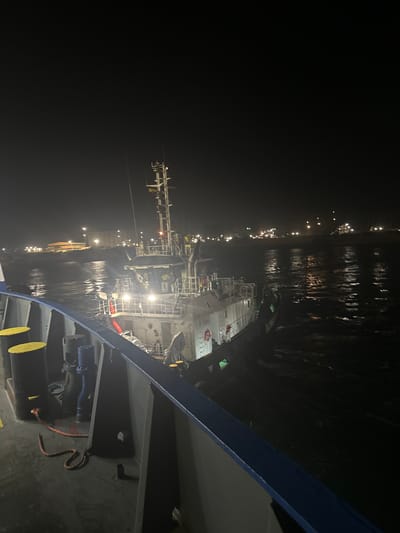 The image depicts a nighttime scene of two boats on the water. The perspective is from the deck of one boat, looking towards another vessel. The second boat is a medium-sized vessel with a superstructure, lights, and various equipment visible. It appears to be docked or maneuvering near the shore, where lights from buildings and infrastructure are visible in the background. The water is dark and reflective, with small waves. The deck of the foreground boat has yellow bollards and a red rope. The scene is likely at a port or harbor. There are no people clearly visible in the image. The location is the city of Uribia, Colombia.