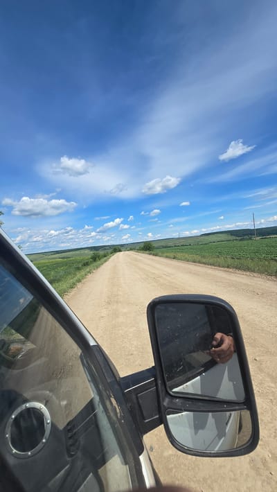 The image shows a view from inside a vehicle looking out at a dirt road stretching into the distance. The road is bordered by green fields on either side, with rolling hills and scattered trees in the background. The sky is blue with scattered white clouds. A side-view mirror of the vehicle is visible in the foreground, reflecting a hand holding a small object. The overall scene suggests a rural landscape in Derevnya Yekaterinovka, Russia, on a sunny day.