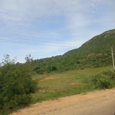 The image depicts a natural landscape with a prominent green hill covered in vegetation and rocks under a partly cloudy sky. The foreground shows a dirt road or path with some scattered green plants and bushes. Several dark power lines stretch across the upper portion of the image. There are no people visible in the scene. The environment appears to be rural or undeveloped, possibly on the outskirts of a settlement. This scene is located in the city of Dorowa, Nigeria.