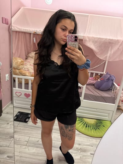 In a dimly lit bedroom with pink walls, a young woman stands in front of a large mirror, taking a selfie. She has dark, wavy hair and is wearing a black satin camisole and shorts. She holds a phone with a patterned case in her right hand. Reflected in the mirror, a child lies in a white crib with pink accents. The crib has a sheer pink canopy. Next to the crib is a large teddy bear. On the floor, a digital scale is visible near the woman's feet, and a kiwi-shaped rug is by the crib. The room appears to be in a private residence in Oreshak, Bulgaria.