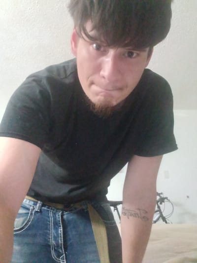 A man is bending forward towards the camera. He has dark hair, a goatee, and is wearing a black t-shirt and blue jeans with white stitching. There is a tan belt around his waist. A tattoo is visible on his left forearm. In the background, there is a bicycle and a white wall. The image appears to be taken indoors. There is no discernible information in the image that indicates its location in Fort Smith, United States of America.