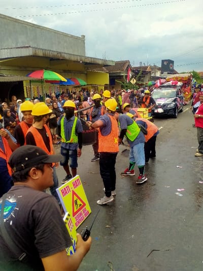 The image depicts a public street scene in Mojogedang, Indonesia, appearing to be a parade or community event under an overcast sky.
In the foreground, a male individual is seen from behind, wearing a black baseball cap and a dark t-shirt. He holds a bright yellow triangular sign with black text reading "HATI - HATI !!!" (Be Careful !!!) and "SEDANG ADA RENOVASI IMAN" (Faith Renovation Underway), accompanied by a black silhouette of a person shoveling. He also holds a black two-way radio in his left hand.
In the midground, a group of approximately seven individuals are present on the wet asphalt road. All are wearing yellow hard hats. Some wear orange high-visibility vests, while others wear neon green vests, over dark shirts, shorts, or pants. One individual in an orange vest is actively striking a black cylindrical object, resembling a drum, with a stick. Two other individuals, one in an orange vest and another in a neon green vest, are bent over, appearing to sweep or shovel scattered debris on the wet road, forming small piles. Other group members stand nearby, some holding tools resembling brooms. One individual wears a black face covering.
The road surface is visibly wet and covered with scattered pieces of colorful paper and other small debris.
In the mid-to-background on the right, a black SUV, possibly a Mitsubishi Outlander, proceeds along the street. It is decorated with pink and white elements on its roof and hood, and appears to be connected to or leading a larger, brightly decorated float, partially visible behind it.
On the left side of the street, a dense crowd of spectators, comprising adults and children, lines the sidewalk. Many female spectators are wearing headscarves (hijabs). Behind the crowd, a two-story building with a light grey upper facade and a yellow lower section is visible. Below its overhang, several brightly colored market umbrellas (red, yellow, green, blue) shade vendor stalls displaying packaged goods. Further down the street, additional buildings are discernible.