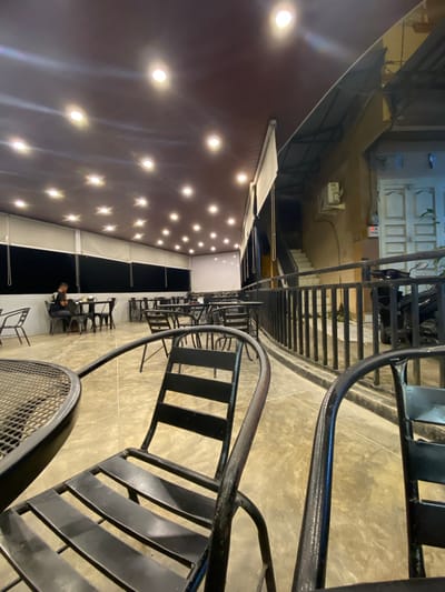 This image depicts an outdoor seating area of a cafe or restaurant, possibly located in Keudeublang, Indonesia, given the context provided. The scene is illuminated by numerous ceiling lights, creating a warm ambiance. In the foreground, two black metal chairs with a distinctive curved back are visible, one partially obscured by the other. A round, black metal mesh table is also in the foreground. Further into the space, several other black metal tables and chairs are arranged, some occupied by patrons. One person is seated at a table in the mid-ground, facing away from the camera. Beyond the seating area, a black metal railing separates the space from a set of stairs leading upwards, and a light-colored building with a closed door is visible to the right. A parked motorcycle is positioned near the stairs. The floor appears to be concrete. The overall impression is that of a commercial establishment with outdoor dining facilities.