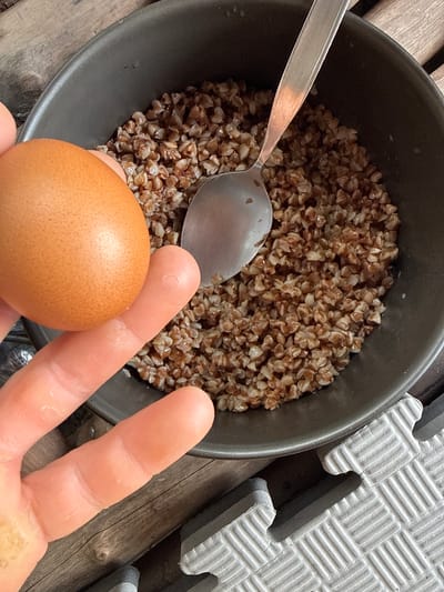A close-up photograph shows a bowl of cooked buckwheat next to an egg held in a person's hand. A silver spoon is partially submerged in the buckwheat. The buckwheat is brown and appears slightly moist. The egg is brown with a smooth surface. The person's hand is pale, with visible fingerprints. The background consists of what appears to be a wooden surface and a gray interlocking foam tile. There is no indication of the specific location in Moo 7, Thailand.