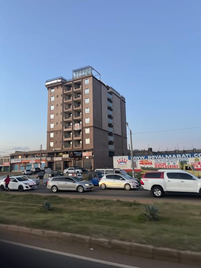 This image displays an urban street scene in Biashara ward, Kenya, under a clear, light blue sky. A prominent ten-story building, labeled "HOMETOWN Apts" at its apex, stands in the mid-ground. This building features a brown facade, numerous white-framed windows, and balconies with glass railings on its right side. A glass-enclosed structure tops the building. Its ground floor houses businesses, including "MAMY TRADERS CENTRE" and "PREMIER OPTICALS," with "TO LET" banners affixed to upper floors. To the left, a sequence of lower-rise commercial buildings is visible. To the right of the main building, a large billboard advertises "ROYAL MABATI.COM" and includes contact information. A multi-lane road in the foreground exhibits moderate traffic. Vehicles visible include a white hatchback with a pink hood stripe, a silver station wagon, a white hatchback, a white pickup truck with a canopy, and a blue tuk-tuk. A grassy median separates the road from the camera's perspective. One individual in a red shirt is standing adjacent to the white hatchback on the far left. A street light pole is visible on the right side of the main building.