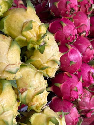 A close-up, overhead shot captures a pile of dragon fruits in what appears to be a market stall in Rai Mai, Thailand. The fruits are a mix of bright yellow and deep pink, with their characteristic green, scale-like skin. The yellow dragon fruits are more prominently displayed in the foreground and to the left of the image, while the pink ones are visible in the upper right portion. The fruits are piled on top of each other, suggesting they are being sold. The lighting is natural and highlights the textures of the fruits. There are no people or any other objects clearly identifiable in the image.