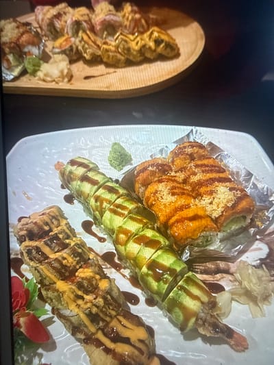 The image shows a close-up of sushi on two different platters. One platter is a white, rectangular dish containing three types of sushi: one with orange topping, a roll wrapped in sliced cucumber and topped with sauce, and a fried roll with sauce. The white platter also includes wasabi and pickled ginger. The other platter is a light brown, rounded-edged wooden board with a single roll of fried sushi and sauce. The setting seems to be a restaurant or dining area. There is no indication of the city of Des Plaines, United States.