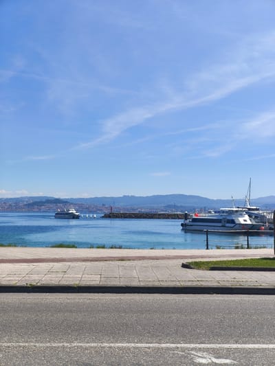 The image depicts a coastal view in Cangas de Morrazo, Spain, under a bright blue sky with thin cirrus clouds. The foreground shows an asphalt road marked with a white line, followed by a gray tiled sidewalk. Beyond the sidewalk, there is a narrow strip of green vegetation before the shoreline.
The water is a clear turquoise blue, stretching to the horizon where a distant cityscape lies against rolling hills. Two white ferry boats are visible: one docked at a pier to the right and the other sailing towards the city. A small red lighthouse stands on a breakwater in the middle distance. A metal railing is visible along the shoreline, acting as a barrier.