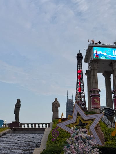 The image captures a scene in Nanshan District, China. Prominently featured is a scaled replica of the Eiffel Tower with red neon Chinese characters along its structure. The tower is situated in front of a screen and architectural columns. To the right of the tower is a large star-shaped decoration and flowering plants. A walkway features two statues along the top, with a water feature along the side. In the background, some additional buildings are visible. The sky takes up the upper left half of the image.
