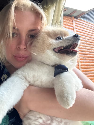 A close-up, selfie-style shot shows a fair-skinned woman with blonde hair holding a small, fluffy white and tan dog. The woman is looking at the camera, with her face partially obscured by her hair. The dog, a Pomeranian breed, is looking up and to the right with its mouth open, panting. It is wearing a dark blue collar. The background consists of a blurred outdoor scene with wooden fencing and some curtains. The image appears to have been taken outdoors in a city.