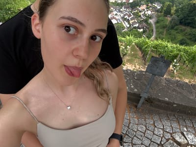 The media file shows a young woman from a low angle, with the background upside down, suggesting a selfie taken from an elevated position. She is wearing a light-colored tank top with thin straps and a delicate necklace with a small diamond pendant. Her blonde hair is visible, with a strand falling near her face. She is sticking her tongue out playfully towards the camera. Her eyes are open and looking towards the lens, with a slight upward gaze.
Behind her, the scene reveals a European-style village with houses clustered together and a winding road. Lush green trees and vegetation surround the village, indicating a natural, possibly hilly landscape. In the foreground, there is a textured stone surface, resembling a cobblestone path or wall, and a metal post is visible. The overall impression is a casual and cheerful moment captured outdoors. The lighting suggests daytime, and the greenery implies it's a warm season, possibly spring or summer. The location context indicates Gernsbach, Germany, which aligns with the architectural style of the village. No visible text is present in the image.