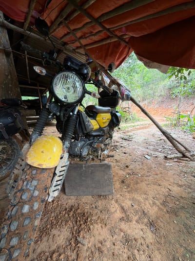 The image depicts a motorcycle parked under a rustic, makeshift shelter constructed from rough-hewn wooden beams and a reddish-orange tarp. The motorcycle is primarily yellow and black, with visible wear and tear, particularly on its tires and front fender which are coated in mud. The ground around the motorcycle is also dirt and mud, suggesting an outdoor, possibly rural, environment. Behind the shelter, dense green foliage and a reddish dirt path are visible, indicating a natural, possibly forested or jungle-like setting. The overall impression is one of a functional but weathered motorcycle in a secluded outdoor location.