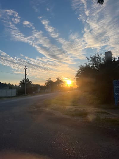 The image depicts a rural road at sunrise in Slobozia, Moldova. The sky is a vibrant blue with streaks of white clouds, illuminated by the bright orange sun low on the horizon. The sun's rays create a hazy glow, casting long shadows across the asphalt road, which appears to be paved and in decent condition.
To the left of the road, a grey concrete fence runs along the property line of a building. Further down the road, a utility pole with wires is visible, and beyond that, trees and residential structures. A white car is parked on the right side of the road, facing away from the viewer.
The right side of the image features a dense cluster of trees, their foliage dark against the bright sky. A portion of a brick structure, possibly a chimney or part of a building, is visible at the top right. The foreground of the road is textured with small pebbles and patches of dry grass. The overall scene conveys a sense of tranquility and the early start of a day in a village setting.