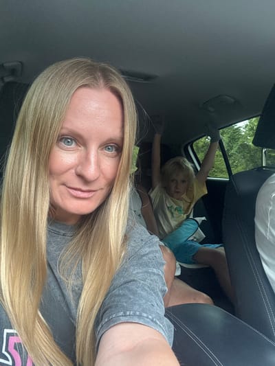 A selfie taken inside a car shows a woman with long blonde hair and blue eyes in the foreground, smiling at the camera. She is wearing a gray t-shirt. In the background, a young child with blonde hair is sitting in the back seat with their arms raised, wearing a yellow t-shirt and blue shorts. The car interior is visible, including car seats and the window showing trees and foliage outside, suggesting a rural or natural setting near Šavnik, Montenegro. The overall scene conveys a casual, everyday moment during a car journey.