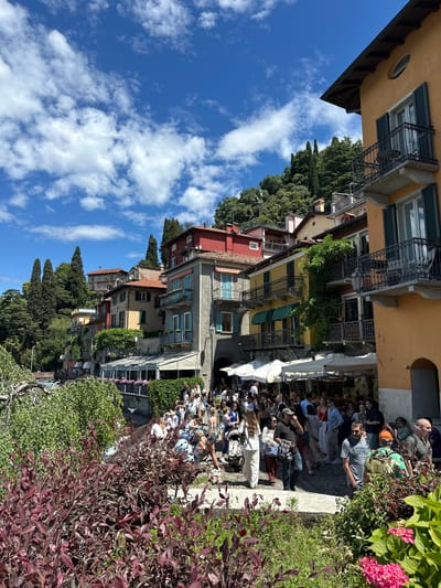 This image captures a vibrant street scene in Varenna, Italy, bathed in bright daylight under a clear blue sky adorned with scattered white clouds.
In the foreground, a lush embankment covered with rich green and reddish-purple bushes, along with some pink flowering plants, creates a natural barrier. Below this, a bustling outdoor cafe or restaurant area is visible, teeming with people. Numerous individuals are seated at tables or standing, socializing and enjoying themselves in casual summer attire. White and green awnings provide shade over parts of the seating area.
The midground and background showcase a charming row of multi-story buildings, typical of Italian architecture. Their facades display a palette of ochre, red, and muted greyish-green. These structures feature balconies with intricate wrought-iron railings, and windows are fitted with dark green or light blue shutters. Tall, slender cypress trees and other dense foliage intersperse with the buildings, cascading down what appears to be a hillside.
The scene suggests a lively atmosphere on a sunny day, likely during the mid-morning or afternoon. The people appear relaxed and engaged in conversation. The architectural style and the abundance of greenery strongly evoke the picturesque charm of an Italian lakeside town. No readable text is prominently visible.