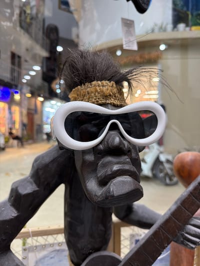 The image depicts a dark wooden statue of a person wearing white sunglasses with a distinctive figure-eight shape. The statue has a headdress of coarse, natural material with dark feathers or strands sticking out. The statue is holding a musical instrument, possibly a guitar or lute, with a dark wooden neck and a visible grid pattern on the fretboard. The background suggests an indoor setting, likely a shop or display case, given the blurred view of shelves stocked with items and bright lights. There is a reflection of the surrounding environment visible on the glass of the display case. There is no indication or evidence to definitively place the scene in Maafushi, Maldives.