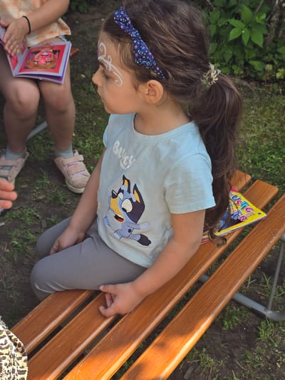 A young girl with face paint sits on a wooden park bench in an outdoor setting, likely in Zirndorf, Germany. The girl has brown hair styled with a floral headband and a clear hair tie. She wears a light blue t-shirt with a cartoon dog design and gray leggings. She is looking down, possibly at a book being held by another person whose legs and pink sneakers are visible in the lower left corner. A children's book rests on the bench next to her. The bench is made of wooden slats. Green foliage is visible in the background.