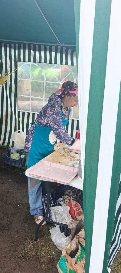A woman stands behind a table at what appears to be an outdoor market or fair, set up under a green and white striped tent. The woman is wearing a patterned long-sleeved shirt, a bright teal apron, blue jeans, and a pink headscarf with a floral pattern. She is also adorned with a beaded necklace featuring metallic coins. She is actively engaged in preparing food, specifically working with small, dough-like pieces laid out on a wooden cutting board. A pink plastic tray with white powder, likely flour, sits in front of her on the table, and more of the dough pieces are visible on the board.
The setting is outdoors, with the ground appearing muddy and covered in patches of grass. To the left of the woman, a blue crate holds some leafy green vegetables, possibly cabbage, and a white plastic bag. Underneath the table, several bags and what look like trash are piled up. One bag is red with white Cyrillic text that appears to read "УГОЛЬ" (UGOL), meaning "coal." Another bag is brown and green with more Cyrillic text, partially visible.
The lighting suggests it could be daytime, possibly overcast, as there are no strong shadows. The overall mood seems to be one of work and preparation, with the woman focused on her task. The striped tent provides shelter, and the setting suggests a rustic or informal event. The location context of Старые Быги, Russia, implies a local community gathering.