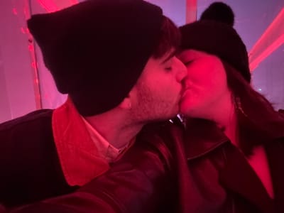 A man and a woman, both wearing black hats, are kissing in a dimly lit space illuminated with red light. The man has short, light-colored hair and a visible beard. He's wearing a jacket with an upturned collar and a partially visible peach-colored shirt. The woman's hair is dark. They are closely embraced, with their faces touching. The background appears to be a room with vertical structures lit in red.