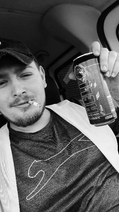 A black and white, eye-level photograph depicts an adult male seated inside a vehicle, likely in the city of Senec, Slovakia. The subject, positioned centrally, wears a dark baseball cap, a dark T-shirt with a light-outlined feline graphic, and a light-colored garment over his left shoulder, secured by a seatbelt extending across his chest. He has a beard and mustache. A lit cigarette is held between his lips. His right hand is prominently displayed, gripping a "HELL Energy Drink Classic" can, which features a red and black design with white text and a stylized devil/demon logo. The background shows the vehicle's interior, including a textured ceiling and a window frame on the right. The subject looks directly towards the camera with a slight smile.