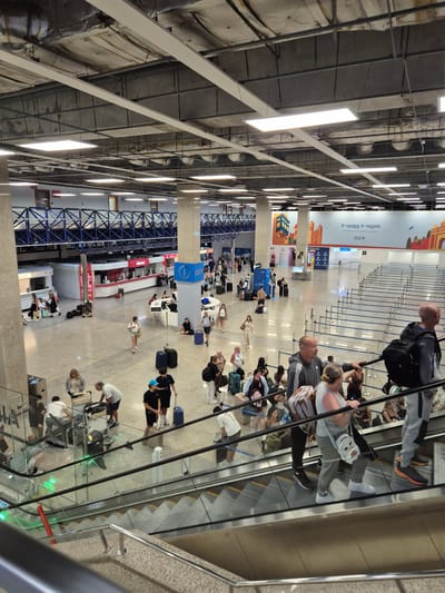 The image depicts the interior of a modern airport terminal in Luqa, Malta. In the foreground, an escalator is visible with several people ascending it. One woman in the foreground is wearing a grey sleeveless top, grey pants, and white sneakers, carrying a backpack. A man behind her is wearing a grey hoodie and black pants. In the middle ground, numerous travelers are seen walking, standing, and sitting, some with luggage. Check-in counters and waiting areas with blue rope barriers are in the background. A large banner with "Il-vjaġġ it-tajjeb" and "Malta Airport" is displayed on the wall. The environment is well-lit with overhead fluorescent lights and appears to be a busy transportation hub.