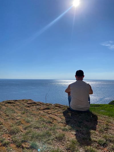 A person wearing a grey t-shirt and a black hat is standing on a cliff overlooking a large body of water under a clear blue sky with the sun shining brightly on the left. The cliff edge is composed of reddish-brown rock and earth with patches of green grass. The location is Шуран, Russia.