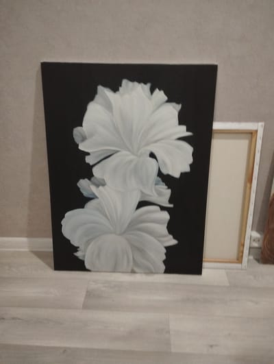 The image displays a vertical painting featuring two large, white, abstract flowers against a black background. The flowers are rendered with soft, rounded petals and a sense of depth. The painting is positioned on a light-colored wooden floor, leaning against a textured beige wall. To the right of the main painting, another canvas is visible, framed in white wood with a beige surface. The overall scene appears to be an indoor setting, possibly a room in a home or studio. No people are present in the image. The location of Balakovo, Russia is not discernible from the visual information provided in the image.