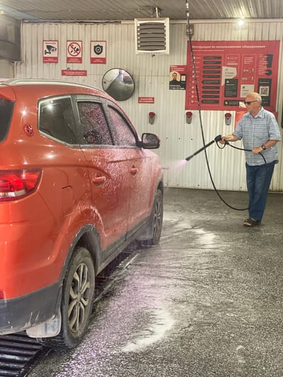 In Novokuznetsk, Russia, a man is operating a high-pressure washer at a self-service car wash. The man, wearing a blue and white striped short-sleeved shirt, blue jeans, and dark sandals, is standing on a wet asphalt surface and directing a pink foam spray towards the side of a red SUV. The SUV is covered in foam and water droplets. The car wash bay features white paneled walls with various signs, including instructions for equipment usage and warning signs about surveillance cameras and security. A large red banner displaying instructions and advertisements is mounted on the back wall. The environment is damp, with foam and water accumulating on the ground.