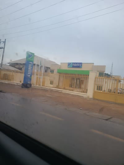 The image shows a Fidelity Bank branch in Karu, Nigeria. The bank building is painted in white and green, with a large yellow metal gate. A prominent blue and green Fidelity Bank sign is visible in front of the building. The road in front of the bank appears wet, suggesting it may have recently rained. The sky is overcast, indicating a cloudy day. The scene is likely during daytime, given the lighting. The image captures the exterior of the bank and its surroundings, which include a street and possibly other buildings in the background. There are no people clearly visible in the frame. The overall impression is that of a typical urban commercial setting in Nigeria.