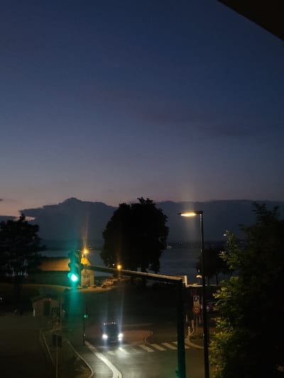 Night scene featuring a street with a lit streetlight and a green traffic light. A car approaches the intersection. In the distance is a body of water with mountains obscured by haze. Trees frame the right side of the image. The sky transitions from dark blue to a lighter gradient towards the horizon. A pedestrian crosswalk is visible on the right.