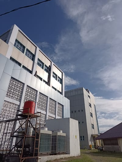 A low-angle, wide shot captures the exterior of two modern buildings against a bright blue sky with scattered white clouds. The building on the left is a multi-story structure with a light-colored facade, featuring decorative geometric patterns on its lower levels and bands of windows on the upper floors. A red water tank is mounted on a metal scaffolding structure to the left of this building, adjacent to several large air conditioning units. To the right of the first building, a second, taller, narrower building with a grey facade and vertical rows of windows stands. In the foreground, to the right of the AC units, a small, dark-tiled roof structure is visible, with a yellow trash bin and a parked scooter near its entrance. The ground is covered in patchy green grass. A dark wire is visible stretched across the top of the frame. The scene appears to be an urban or institutional setting within the city of Mesjid Reuntoh, Indonesia.