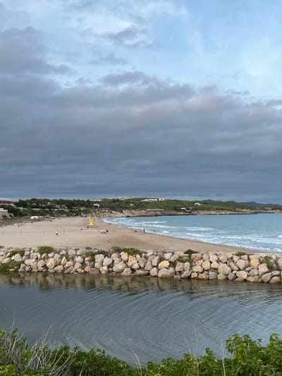 The image captures a coastal landscape at Platja de l'Aiguadolç in Vilanova I La Geltrú, Spain, under an overcast sky. In the foreground, a calm, dark-toned body of water, likely a river or canal mouth, occupies the lower left. This water is separated from a sandy beach by a rock breakwater constructed from large, light-colored stones. Sparse green vegetation grows along the immediate edge of the foreground water and the base of the breakwater.
The midground features a broad, light-brown sandy beach curving gently from left to right. The Mediterranean Sea extends from the shoreline to the horizon, exhibiting light blue-green water with gentle breaking waves. Approximately eight individuals are scattered across the beach; a group of three to four people stands near the water's edge on the right, while other figures are individually distributed further along the sand. A yellow cylindrical structure, consistent with a lifeguard station, is positioned centrally on the beach. Two wooden poles, indicative of a volleyball net, are visible on the far left of the beach.
The background consists of rolling green hills densely covered with trees, rising behind the beach and coastline on the right. Several white structures, possibly residential buildings, are discernible amidst the foliage. The sky is predominantly covered with layered grey clouds, with lighter blue areas visible in the uppermost portion. The overall lighting is diffused.
