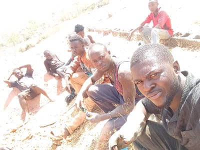 A group of seven men are posing outdoors in a field or dirt area under bright sunlight. Most are looking at the camera and some are holding items. One man wears a black shirt and has a beard. Several men are wearing tank tops and casual shirts. The ground is dry with sparse vegetation. The image is slightly overexposed, particularly on the ground.
