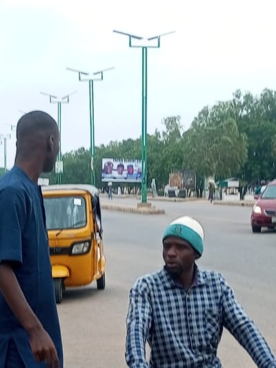 In Wuro Dole, Nigeria, a man in a blue traditional outfit stands on the left, his back to the camera, looking towards the right. In the center of the frame, a yellow auto-rickshaw is parked, its doors slightly ajar. To the right of the auto-rickshaw, another man wearing a blue and white checkered shirt and a teal and white beanie is in motion, possibly riding a bicycle, looking to his left. In the background, a red car is visible driving on the road, with trees and a billboard displaying images of what appear to be ceramic items lining the street. Several solar-powered streetlights are prominently featured throughout the scene. The ground is paved, suggesting an urban or semi-urban environment.