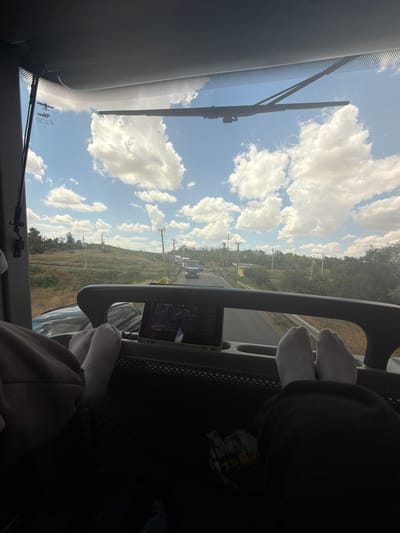 The image depicts the view from the interior of a vehicle, looking out through the windshield. Two pairs of feet, clad in white socks, are prominently visible in the foreground, resting on what appears to be the dashboard or a similar surface. One set of feet is positioned on the left side of the frame, partially obscured by dark clothing, while the other set is on the right. Between the pairs of feet, a tablet device is mounted, displaying a dark image, possibly a video.

The windshield offers a view of a road stretching into the distance, flanked by dry, grassy terrain and sparse trees. Utility poles line the side of the road. A convoy of trucks is visible on the road ahead. The sky is blue with scattered, fluffy white clouds, suggesting daylight and fair weather. A windshield wiper is positioned across the top of the windshield. The location is specified as Kyrnychky, Ukraine.