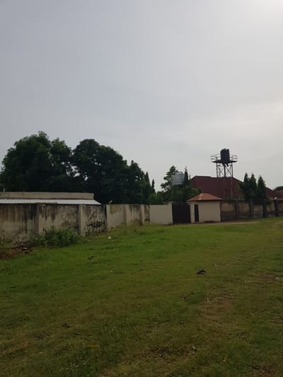 The image displays an outdoor scene with a focus on a grassy field adjacent to a low concrete wall. Beyond the wall, there are buildings with reddish-brown roofs and some trees. A prominent black water tank is visible on a metal structure atop one of the buildings. The sky is overcast, suggesting a cloudy or hazy day. The overall setting appears to be a somewhat industrial or residential area, possibly within a compound given the presence of the wall and buildings. There is no visible text in the image. The perspective is somewhat tilted, giving a slightly unusual orientation to the scene. The lighting suggests it could be daytime, but the overcast conditions obscure any strong shadows. The grass is green, indicating it's likely not a dry season.
