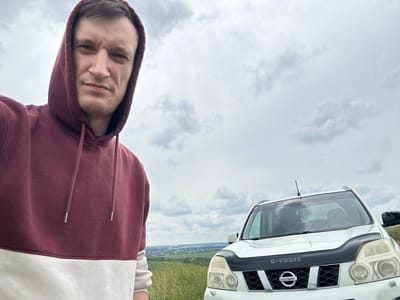 A man wearing a maroon and white hooded sweatshirt stands in a field, looking directly at the camera. Behind him, to the right, is a white Nissan X-Trail SUV. The vehicle has a black hood deflector and a black grille with the Nissan logo. In the background, a city skyline is visible in the distance, with green fields and tall grass in the foreground. The sky is overcast with scattered clouds. This scene is located in the city of Humentsi, Ukraine.