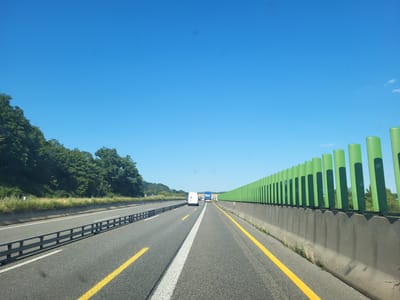 The image depicts a view from a vehicle driving on a highway on a clear, sunny day. The sky is a vibrant blue, suggesting it is daytime, likely late morning or afternoon.

The highway is asphalt with distinct yellow and white lane markings. On the left side of the road, a metal guardrail separates the lanes from a grassy embankment and a dense line of green trees. On the right side, a concrete barrier topped with a row of bright green, cylindrical noise reduction elements creates a striking visual contrast against the blue sky.

Several vehicles are visible on the highway. In the distance, a white van and a blue truck with "GLS" branding are traveling in the same direction as the viewer. The presence of these vehicles indicates that this is an active roadway, likely a major route.

The scene appears to be in a rural or semi-rural area, given the presence of trees and open skies. While the exact location is specified as Laasdorf, Germany, no specific landmarks are clearly identifiable from the image itself, other than the general highway infrastructure. The overall mood is serene and open, conveying a sense of travel and a pleasant day for driving. There is no visible text other than the "GLS" logo on the truck.