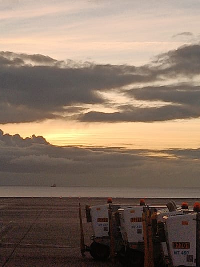 A serene scene unfolds with a layered sky of pink-tinged clouds, transitioning to a band of yellow sunlight. Below, a flat expanse of water separates the sky from the ground, with a distant ship visible on the horizon. In the foreground, several white airport carts with orange lights are clustered, their identification numbers visible. The ground appears to be concrete, adding to the industrial aesthetic.