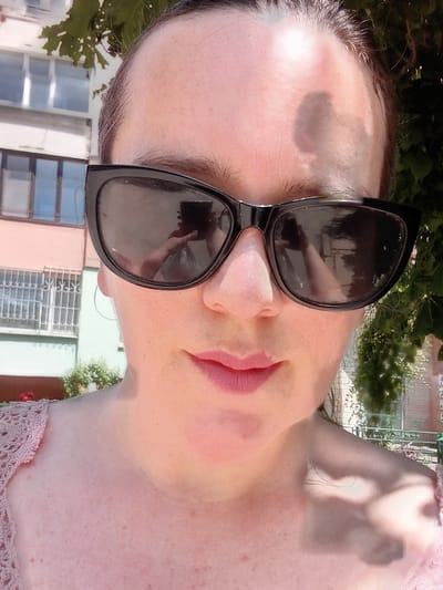 This is an upside-down, close-up media file of a person's face and upper torso, taken outdoors during the day. The subject is wearing dark-rimmed sunglasses with reflective lenses that show a distorted reflection of what appears to be a phone being used to take the photo. The person's lips are painted with pink lipstick. Their skin appears fair with some freckles. The background shows a building with windows and some greenery, suggesting an urban or suburban setting. The overall mood is casual and relaxed. No text is visible in the media file.