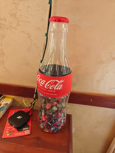 This image depicts a decorative Coca-Cola bottle, filled with bottle caps, standing on a wooden surface. The bottle has a red cap and a red label with the Coca-Cola logo. Next to the bottle, on the same surface, is a black electronic device labeled "A1" and a red promotional flyer. The background features a textured wall. The scene is located in the city of Peshtera, Bulgaria.