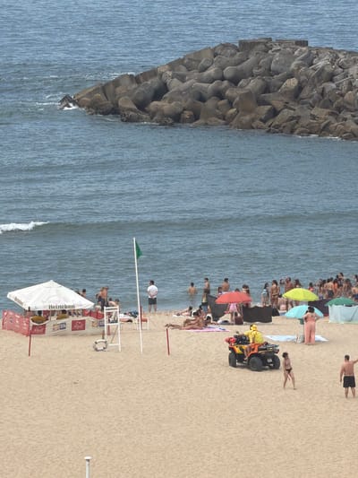 This is an aerial, medium-shot photograph of a crowded beach in Espinho, Portugal, on a hazy day. The scene is dominated by the sandy beach in the foreground, the choppy blue ocean in the midground, and a large, concrete breakwater made of tetrapods in the background. Numerous beachgoers are spread across the sand, many in swimwear, engaging in various activities. Some are lying on towels, others are standing and talking, and a group is partially submerged in the water. Beach umbrellas, a white tent with "Heineken" branding, and a red safety barrier are visible on the sand. A green flag is planted on a pole near the water's edge. In the mid-right of the frame, a yellow ATV with two individuals in yellow vests, likely lifeguards, is parked on the sand. Several beach umbrellas in red, yellow, and green hues are scattered amongst the beachgoers. The breakwater, constructed of numerous interlocking concrete tetrapods, extends into the ocean, protecting the coastline. The overall impression is of a busy summer day at the beach.