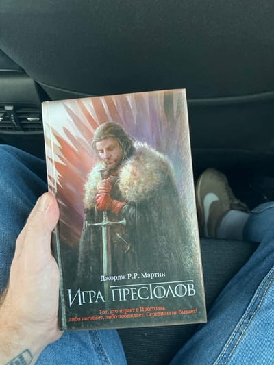 The scene depicts a close-up, downward-angled view inside what appears to be a vehicle in Emek Izrael Regional Council, Israel. The primary subject is a book titled "ИГРА ПРЕСТОЛОВ" (Game of Thrones) by "ДЖОРДЖ Р.Р. МАРТИН" (George R.R. Martin), held upright in the left hand of an individual.
The book's cover features an illustration of a male figure with long, dark hair and a beard, dressed in a dark, fur-trimmed cloak, grasping a two-handed sword with hands protected by red gauntlets. The background of the illustration displays a pattern of vertical, light-colored streaks against a darker, reddish-purple base, suggesting a cold environment. Below the author's name and title, a Russian quote reads: "Тот, кто играет в Престолы, либо погибает, либо побеждает. Середины не бывает!" (He who plays the game of thrones either wins or dies. There is no middle ground!).
The hand holding the book exhibits a light skin tone, and a faded, partially obscured tattoo is visible on the back of the wrist. The individual holding the book is wearing blue denim jeans. To the right, another person's lower leg and foot are visible, also clad in blue denim jeans, a grey sock, and a brown athletic shoe featuring a prominent white Nike swoosh logo on its side. The immediate environment consists of dark, likely upholstered interior surfaces, consistent with a vehicle's seating or dashboard.
