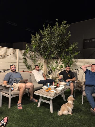 Four men and a dog are gathered in a backyard in Lod, Israel. The scene appears to be an evening gathering. The men are seated on a couch and chairs around a low table. One man on the left is using a laptop and smiling at the camera. Another man next to him is looking away. A third man is sitting in a chair to the right of the table, looking thoughtfully to the side. The fourth man on the far right is leaning back in his chair with his hands behind his head. The table is set with bowls of snacks and drinks. The backyard is enclosed by a white wooden fence and features a tree with green leaves. The ground is covered in artificial grass. The lighting suggests it is nighttime.