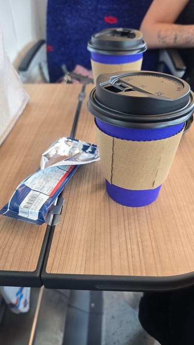Two disposable coffee cups with brown sleeves and blue bottoms are placed on a wooden table. The cups have black lids with a "CAUTION CONTENTS HOT" warning. One cup is closer to the foreground, and its lid is slightly open. A crumpled silver and blue snack wrapper is attached to the edge of the table with a clip. The background shows a person's arm with tattoos and a blue, patterned fabric. The scene appears to be indoors, possibly on a train or in a cafe, given the table and seating arrangement. The text visible on the wrapper includes nutritional information in Hebrew, with dates "05-01-2021" and "03:34", and a barcode "7290019310679". The overall impression is a casual, everyday moment.