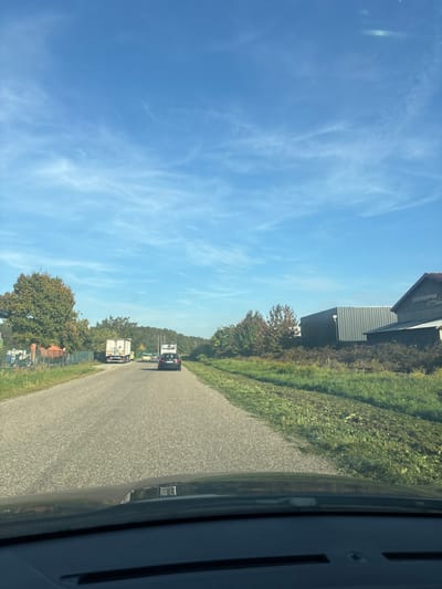 The image displays a daytime scene from the perspective of a vehicle traveling on an asphalt road in Scherwiller, France. The sky is clear blue with sparse, wispy white clouds. Ahead, a dark-colored sedan with a prominent light-colored, rectangular object mounted on its roof is visible, traveling in the same direction. Further down the road on the left, a white semi-trailer truck is present, along with other indistinct vehicles, including one appearing yellow. The left side of the road is bordered by a grassy verge and a line of trees exhibiting both green and autumnal foliage, with some fencing visible among them. The right side of the road features a grassy shoulder with some mown areas, followed by a row of trees with similar mixed foliage. Beyond these trees, two buildings are discernible: a modern-looking structure with grey corrugated metal siding and an older building featuring a red-tiled roof. The dashboard of the observer's vehicle is visible at the bottom of the frame.