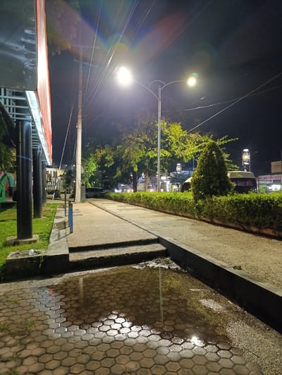 The image shows a sidewalk in Kecamatan Asembagus, Indonesia. The sidewalk is made of concrete and brick pavers, with a small puddle in the center. To the left of the sidewalk, there is a large billboard. To the right, there is a grassy area with a few trees in the background. The street is deserted and there are only a few cars parked on the side of the road. There is a streetlight illuminating the scene. The sky is dark and overcast. The environment is a night scene with streetlights and street signs.