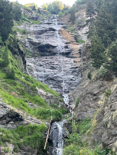The image shows a tall, narrow waterfall cascading down a steep, rocky mountainside covered in patches of green grass and sparse trees. The rocks are layered and appear to be of a dark grey color, with some lighter, tan-colored sections visible higher up the waterfall. The water creates white streaks as it flows down the textured surface, pooling at the bottom before continuing its descent over more rocks and vegetation. The surrounding environment is a rugged, natural landscape, characteristic of a mountain region. There are pine trees visible on the slopes to the left and right of the waterfall. The lighting suggests it is daytime, with sunlight illuminating the upper portions of the scene. The image's perspective is from below, looking upwards at the waterfall and the mountain. This waterfall is located in the vicinity of Benasque, Spain, within a mountainous terrain.