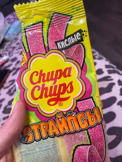 A hand holds a yellow bag of Chupa Chups "Sour Stripes" candy. The bag features a bright yellow background with red and pink candy strips visible. The Chupa Chups logo is prominently displayed in the center. The text on the bag is in Russian, with "КИСЛЫЕ" (kislye - sour) at the top and "СТРАЙПСЫ" (straypsy - stripes) at the bottom. The image shows no discernible connection to Borisova Griva Massiv, Russia, nor does it depict any specific environmental details of that location. The focus is solely on the product packaging.